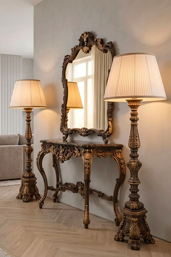 A hand-carved wooden Baroque-style console table with a marble top. Above it is a Baroque-style mirror with an inlaid wooden frame. On either side are standing baroque lamps, also made of carved wood.